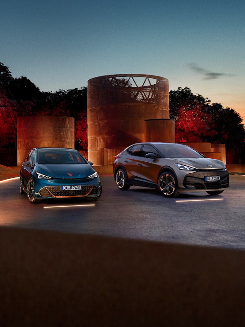 CUPRA Born und CUPRA Tavascan nebeneinander auf einer beleuchteten Fläche vor architektonischem Hintergrund