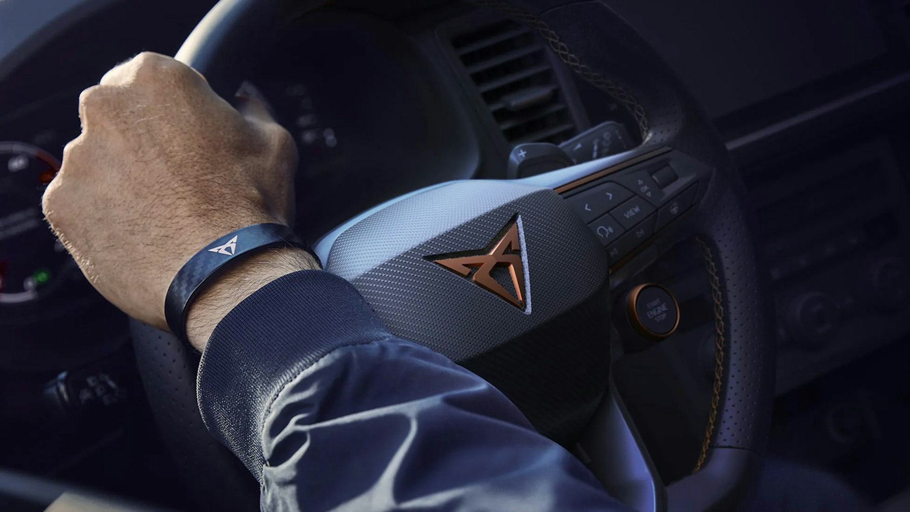 Detailaufnahme eines CUPRA-Lenkrads mit kupferfarbenem Logo, Hand am Steuer mit CUPRA-Armband