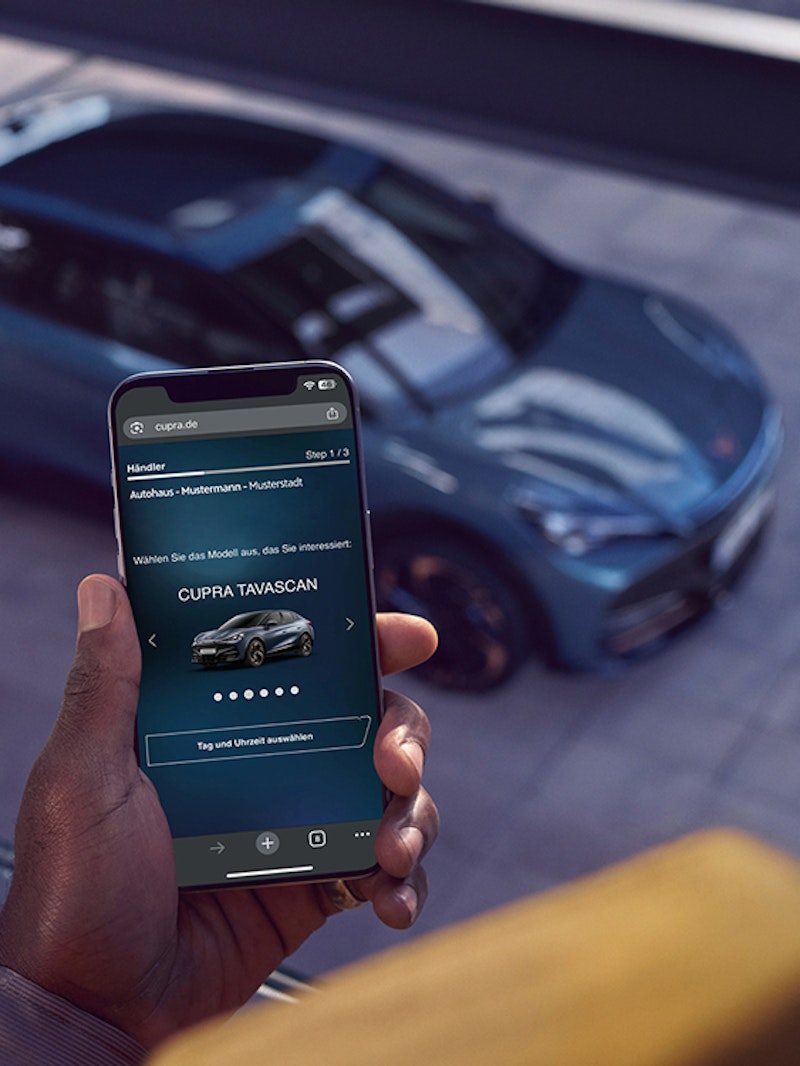 CUPRA Tavascan von schräg oben mit Person im Vordergrund, die eine Probefahrtbuchung am Smartphone tätigt