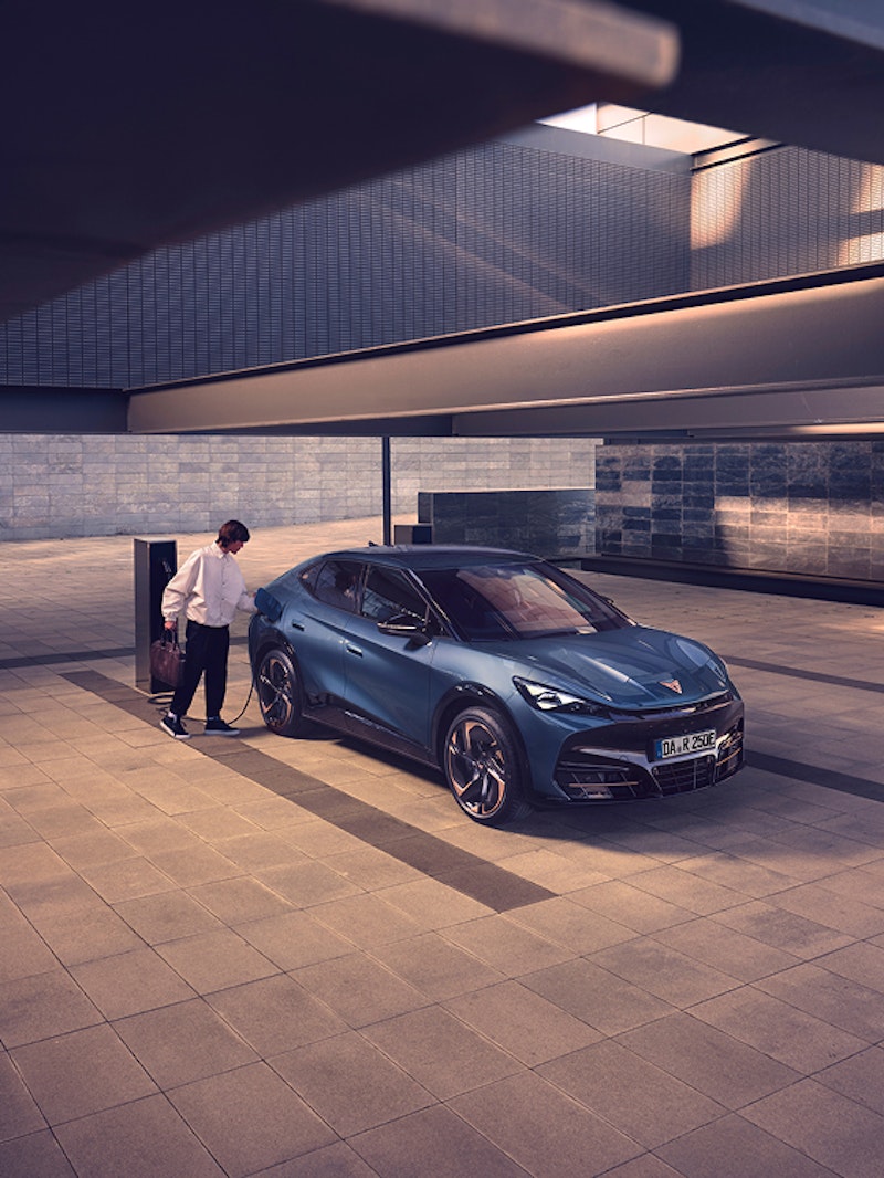 Eine Person steht neben einem CUPRA Tavascan in Blau und lädt das Elektro-SUV an einer Ladesäule in einer urbanen Umgebung auf.