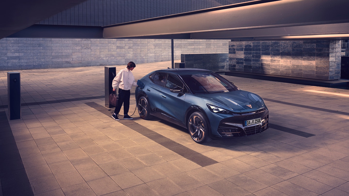 Eine Person steht neben einem CUPRA Tavascan in Blau und lädt das Elektro-SUV an einer Ladesäule in einer urbanen Umgebung auf.