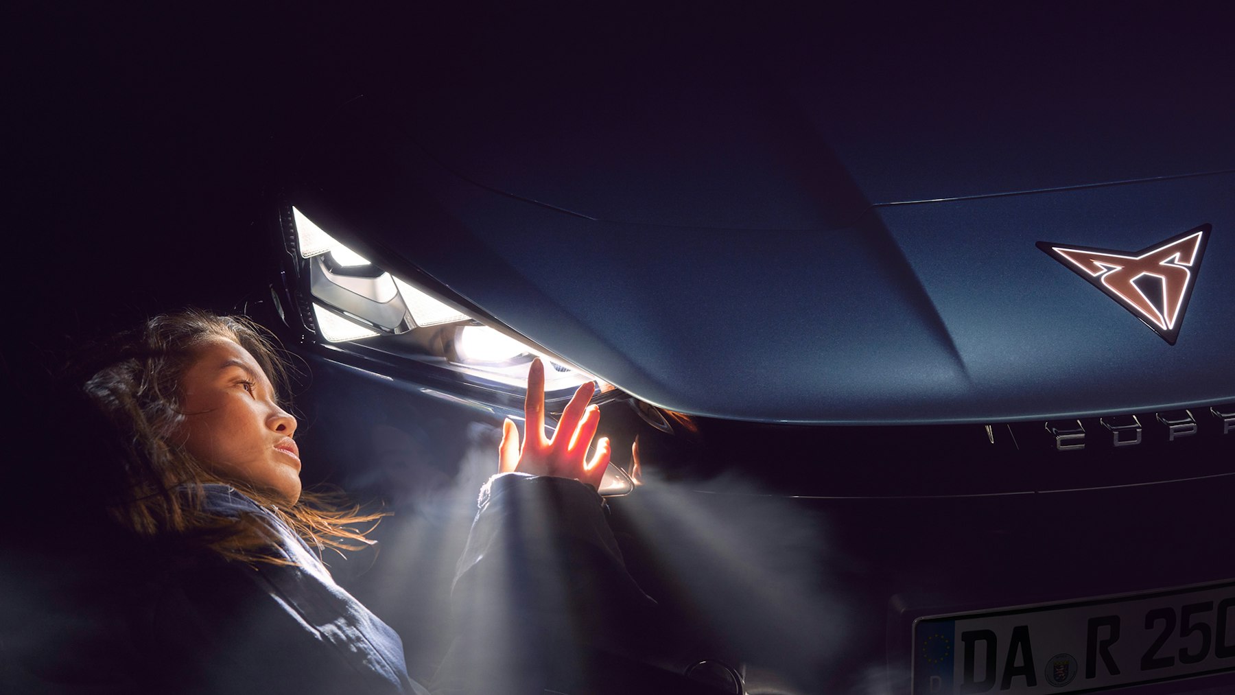 Frau in Nahaufnahme blickt auf den eingeschalteten Frontscheinwerfer eines CUPRA Tavascan, im Vordergrund das beleuchtete CUPRA Logo auf der Motorhaube