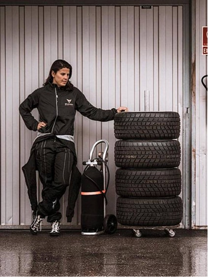 Frau in CUPRA Outfit lehnt an einem Stapel Autoreifen neben einem CUPRA Rennfahrzeug in einer Garage.