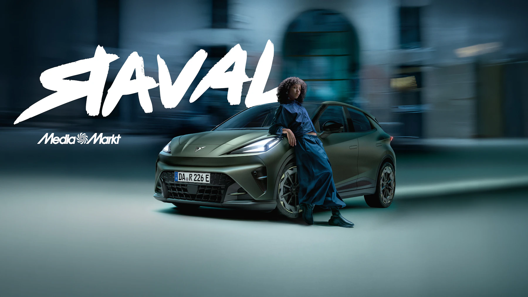 CUPRA Raval in der Lackfarbe Plasma Exclusive in Dreiviertel-Frontansicht, geparkt auf einem naechtlichen Kopfsteinpflaster vor einer Hausfassade mit dem Schriftzug 'Beyond Rebel Raval'.